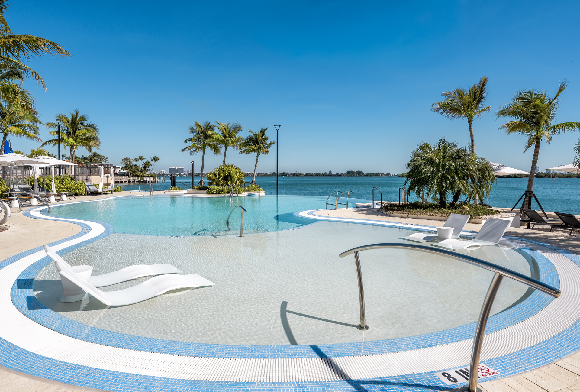 1-Bedroom Luxury Apartments in North Miami, FL - Biscayne Shores - Waterfront Swimming Pool with Lounge Chairs in the Water, Tanning Chairs on the Sand Surrounded by Palm Trees